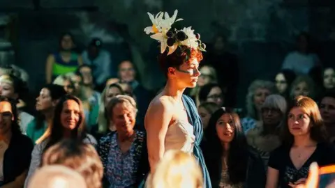 Dave Challenger Photography A woman walking on a catwalk wearing a white and blue dress and white lilies on her head. People are watching from the sides.