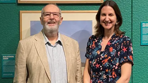 MNH A man wearing a beige blazer and pale shirt standing alongside a woman wearing a black dress with colourful sea creature designs standing in against a green wall with paintings hanging on it. Both are smiling.