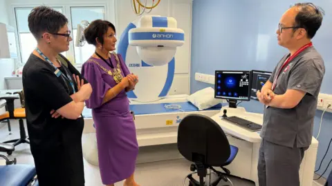 Sheffield Teaching Hospitals NHS Foundation Trust Ms Dunn speaks to a male staff member who stands next to a white desk with two monitors and a keyboard. Another member of staff stands next to Ms Dunn on her right.