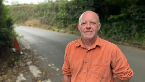 A man in an orange shirt stands in a country lane. There are thick, high hedges behind him. 