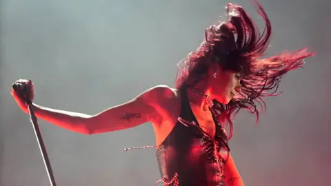 PA Media Dua Lipa on stage at Glastonbury in June. She is wearing a sleeveless top, laced up at the front. Her head is flung to the left with her hair swinging around her head in the movement. Her right arm is held out and holding onto the top of a microphone stand