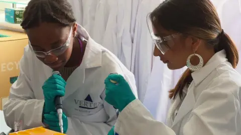 BBC Two teenage women wearing white lab coats, lab glasses and green rubber gloves, using pipettes.