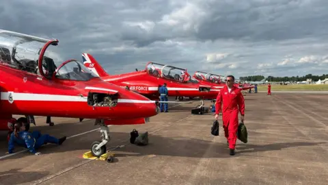 BBC The Red Arrows on the tarmac at RAF Fairford, with a pilot walking in front of them holding two backpacks