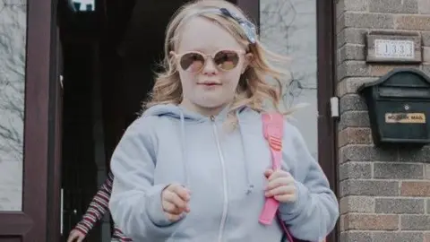 Banjoman Films, supported by Screen Ireland Claudia walking towards the camera wearing a pair of black jeans, a grey zip-up hoodie and white trainers. She has blonde hair, pink sunglasses and a pink backpack. In the background is a house with the front door open.