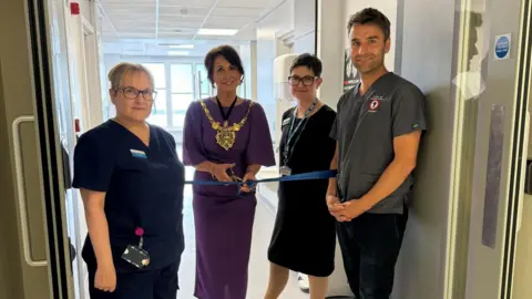 Sheffield Teaching Hospitals NHS Foundation Trust Ms Dunn wears a purple dress as she holds scissors to cut the blue ribbon stretched between two doors. Three trust staff stand next to her, including Dr Ball on the very right. 