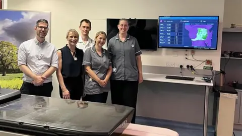 NHS Humber Health Partnership Five members of the hospital's radiotherapy team smliing at the camera standing beside a medical table and a computer screen