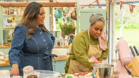 Channel 4/Love Productions/Mark Bourdillon Alison Hammond in a denim dress with curled brown hair is stood on the left. Illyin Morrison is piping some red icing. She is wearing a green jumper and a bakers apron. 