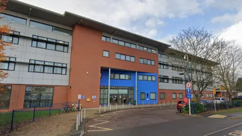 Google The entrance to The Stanway School. It is a large building which is grey, orange and blue in appearance.