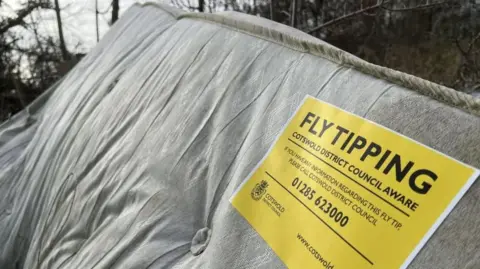 BBC An old mattress dumped near some trees. It has a yellow fly-tipping sticker on it from the council that says 'Fly tipping. Cotswold District Council Aware'. The sticker appeals for anyone with information about the fly-tip to contact them. 