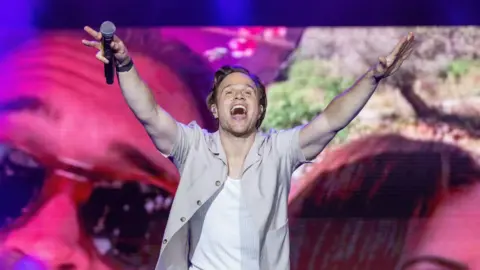 PA Olly Murs performing on stage in a grey open shirt with a white T-shirt underneath. He has is arms outstretched with a mic in one hand and a big smile on his face. 