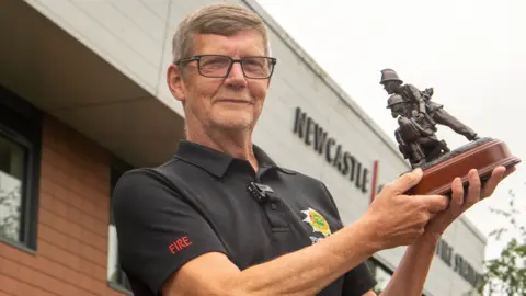 Staffordshire Fire and Rescue Service Kelvin Knapper wearing a black Staffordshire Fire and Rescue Service shirt holds up his award - a trophy featuring two miniature firefighters.