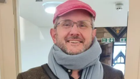 Supplied A smiling man with a beard and glasses wearing a red flat cap and light  blue scarf