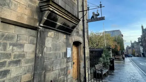 BBC Photo of the building in Edinburgh's tollbooth. It is the side of a blonde sandstone building. It looks old and has a wooden door with iron rivets