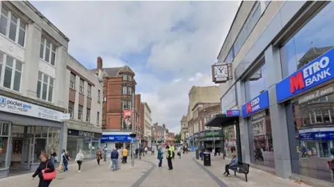 Google A view of a busy shopping street, pedestrians are seen walking throughout, there are rows of shops on either side and to the right is a building with a sign that says Metro Bank