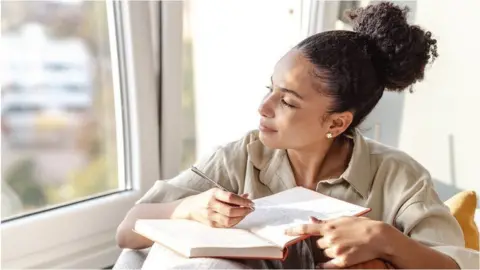Getty Images A young woman sitting on a sofa looking to her right. She has dark hair pulled back into a high ponytail and is wearing a beige short-sleeved shirt. She has a journal open in front of her, leaning on the arm of the sofa. She is holding a pen in her right hand and it is hovering above the page, where writing can be seen
