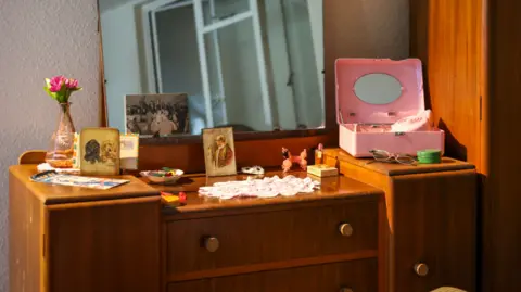 Museum of the Home  A room set in 1956 which has a dressing table on display that has items including a small vase with a pink flower, old photos, a pair of glasses and a pink make up case