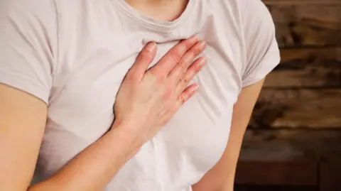 Getty Images A woman is holding her hand on her chest to indicate a problem with her heart. She is wearing a light pink T-shirt
