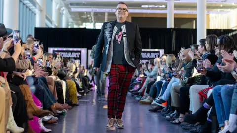 Stuart Trevor Stuart Trevor on the catwalk at one of his shows wearing red and blue check trousers, trainers and blue decorated jacket with an audience sat around him taking photos on their phones