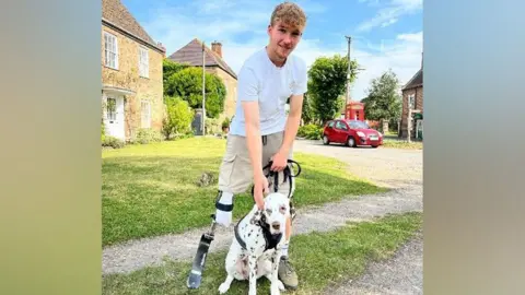 BBC Ollie standing with his prosthetic blade alongside a Dalmatian dog on a lead in a village setting on a sunny day