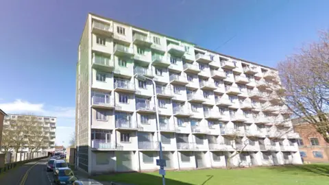 Google A large white block of flats sitting on a green space, surrounded by houses and other blocks of flats. The sky is blue, and cars are parked on the road which runs to the left of the block.
