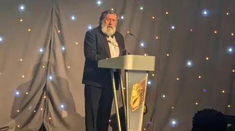 RTS A man in a dinner jacket, white shirt and black bow tie standing behind a black lectern, speaking into a microphone. There are black curtains behind him with multiple spot lights. Tomlinson has a full white beard and long greying black hair.