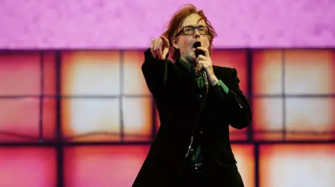PA Media Jarvis Cocker, wearing glasses and a brown jacket, stands on stage pointing his finger and holding a microphone.