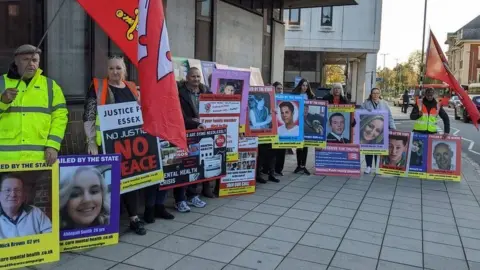 Stuart Woodward/BBC A group of at least 11 campaigners standing outside an inquest in Essex. They are holding up flags and placards with the names and photos of family members who have died. At the top of many placards, it says "failed by the state" in capital letters