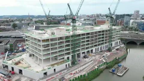 Ross James An aerial image of the new building under construction. The main frame of the building is complete, with three cranes beside the structure. It is a large rectangular building with five floors and is situated next to the river. The skyline landscape of the city can be seen in the background. 