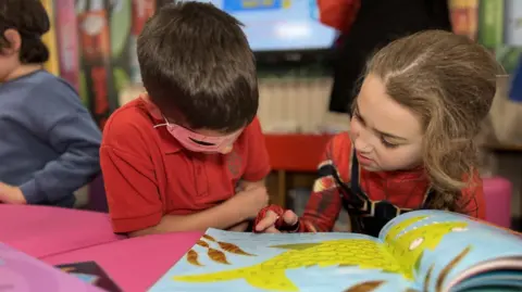BBC A young boy and girl are reading a book. The boy has dark hair and is wearing a Lone Ranger-style mask while the girl has light brown hair and has her finger paused over a word in the book.