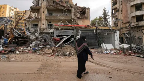 Reuters A woman dressed in a black headscarf and dark clothing walks past the wreckage of heavily damaged buildings in Beirut’s southern suburbs, Lebanon. The scene shows extensive destruction, with collapsed walls, twisted metal and debris scattered across the ground. 
