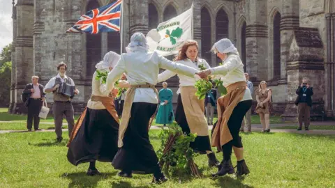 Create Studios People in historical dress dancing in a circle with a village flag and oak branches