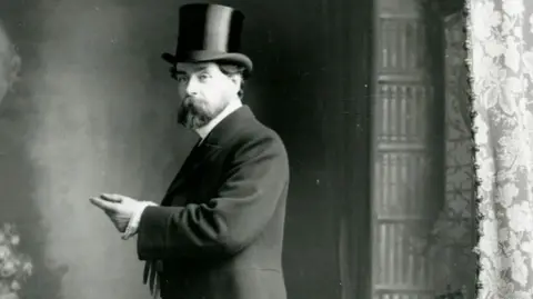 MNH A black and white formal portrait photograph of Archibald Knox, who has a beard and is wearing a formal jacket and shirt with a stiff collar along with a top hat.