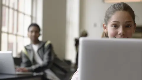 Getty Images A young girl sits at a laptop while a male student, blurred by the camera, sits behind