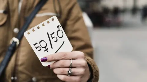 Getty Images A woman in the street shows a paper with the text 50/50 written in it, with the zeros as the female and male gender symbols, depicting the gender parity concept.