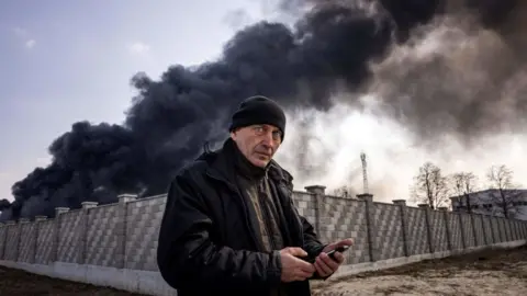 Getty Images/Fadel Senna A man staring at the camera as he holds a phone with black billowing smoke in the background