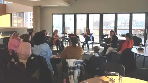 Handout Image of a Mothers in Music session. The group of women are sat in a circle. A few of them can be seen smiling, and Alice can be seen talking, with her arms stretched out.