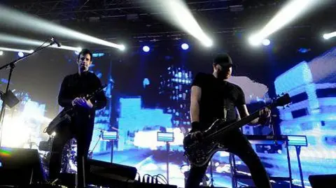 Pendulum on stage- Two men dressed in black with guitars with a blue city skyline background