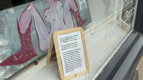 A painting of a naked woman with her legs spread in a gallery window, with a small notice in front of the painting telling visitors they could leave comments in a book.