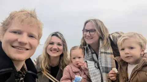 Ed Sheeran Ed Sheeran poses for a selfie with Amanda Jane, her friend, and their children. Ed has ginger hair and is smiling into the camera. Amanda is wearing glasses and a scarf while her female friend has blonde hair. The two children pictured are each holding a piece of food and staring into the camera.
