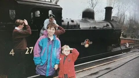 Rare Book Auctions An photograph taken in the 1990s of Christine McCulloch wearing a raincoat and a young Adam wearing a red coat as the pair pose next to a steam train 