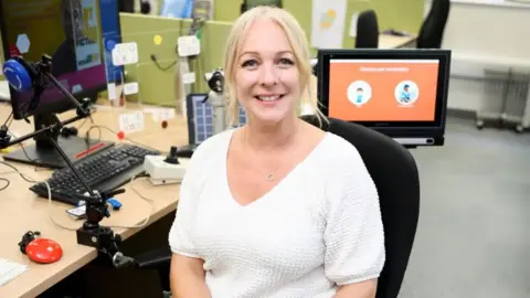Cambridge University Hospitals NHS Foundation Trust Jenny Boucher in a white top with blonde hair, sat on a black chair at a wooden desk 