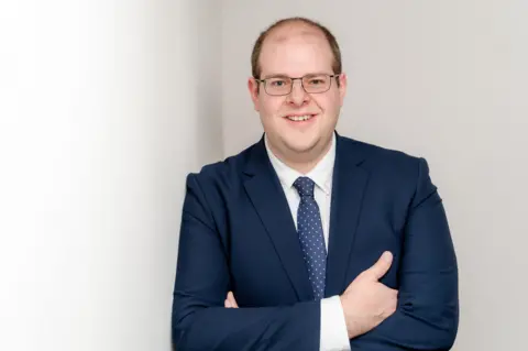 Conservative Party Conservative candidate Jonathan Ash-Edwards in a navy blue suit and tie, wearing glasses