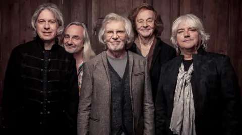 Alex Lake The Zombies band in a recent photo. Rod Argent is centre wearing glasses and a speckled suit jacket and striped T-shirt, he is smiling at the camera and has grey medium-length hair and stubble. 