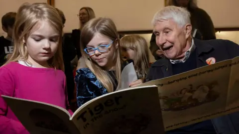 Jamie Simonds Skye (a girl with blonde hair and is wearing a pink top) she is stood next to Célestine (she is wearing a blue velvet top and has blonde hair and blue glasses) she is next to Nick Butterworth who is reading two books.