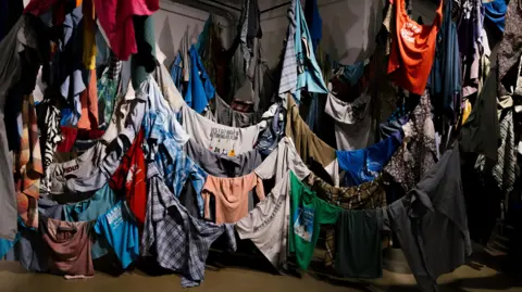 Blake Watson Photography Shirts of different colours, designs and sizes are hung indoors around lines that drop down close to the floor where some of the shirts are touching the floor. There are hundreds of shirts on display.