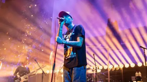 Getty Images A singer on stage, with a bassist to his right. He is wearing jeans, a black printed t-shirt and cap. Tattoos can be seen on his bare arms and the stage is lit with purple and yellow lights.