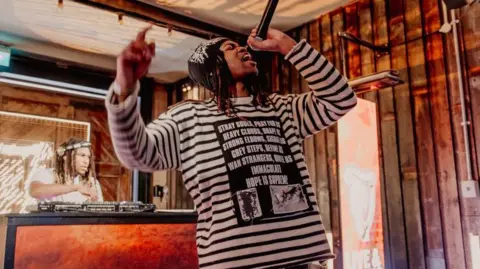 Getty A man in a black and white striped shirt hold a microphone. He has long black hair and has on a beanie. Behind him is a DJ spinning turntables