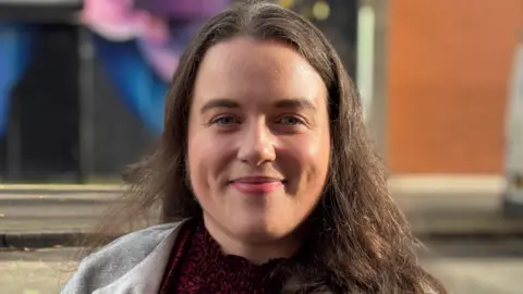 BBC Áine Murray looks at the camera, smiling slightly. She has long, dark hair and is wearing a red patterned top and a light grey top.