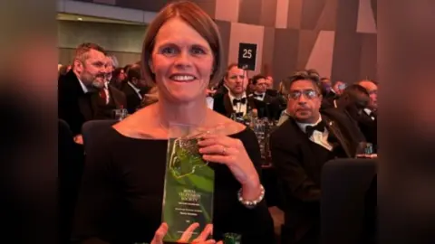 BBC A woman in a black off-the-shoulder dress holding up an oblong glass award. It is engraved with Royal Television Society, then in smaller letters says Midlands Awards 2024, Breakthrough on Screen Unscripted, Nicola Goodwin, BBC. The woman has short brown hair in a bob. Other guests in black tie can be seen at tables behind her in an events venue.