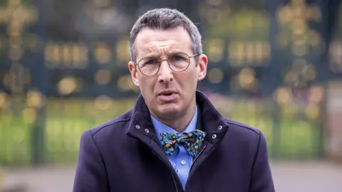 PA Media Andrew Muir looking at the camera. He is pictured from the chest up. He has grey hair and is wearing a blue shirt, a bow tie, navy jacket and round glasses. He has a serious expression on his face. The background of the image is blurred. 